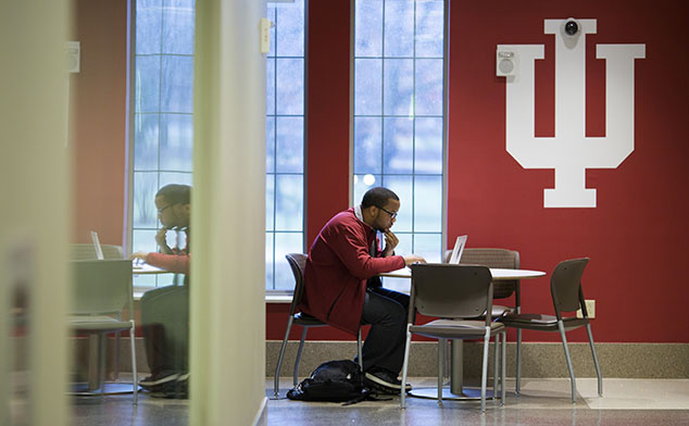 A male student working at a desk on his laptop in front of a big block IU logo on the wall.